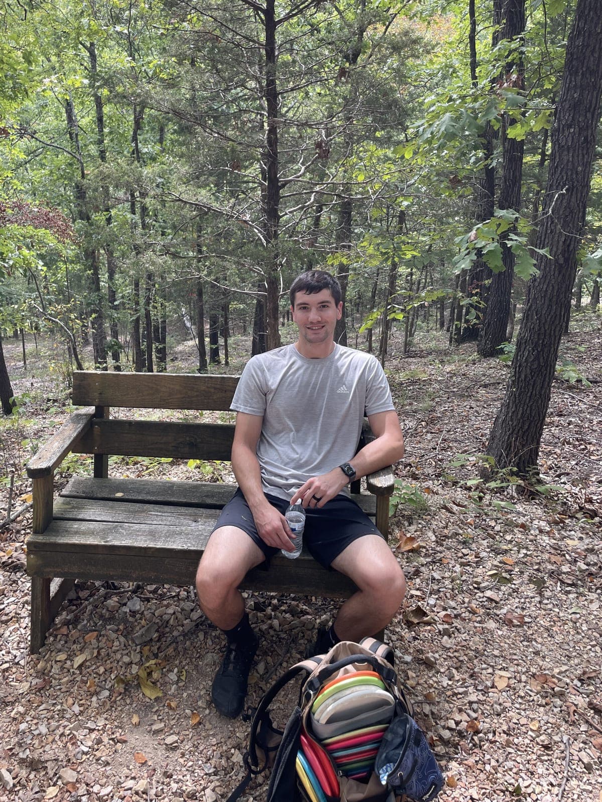 Justin sitting on a bench on a wooded disc golf course, his bag at his feet